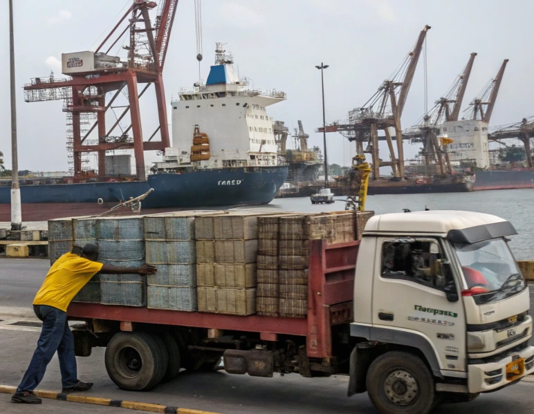 Worker loading cargo onto a truck near container cranes and ships, illustrating ports news impact of labor pressure and slowed automation in port operations
